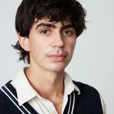 Young man with dark hair wearing a white collar shirt and a black V-neck sweater. 